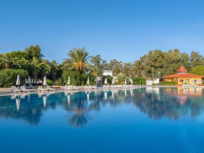 Velký hotelový bazén s lehátky, palmami a jasně modrou oblohou v pozadí.