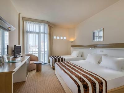 Hotelový pokoj se dvěma manželskými postelemi, psacím stolem, židlí a balkonem se světlými závěsy.