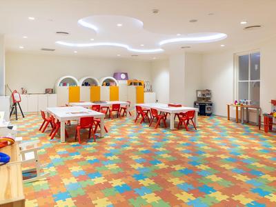 Jasny, duży pokój zabaw dla dzieci z kolorową podłogą i małymi stolikami z czerwonymi krzesłami.