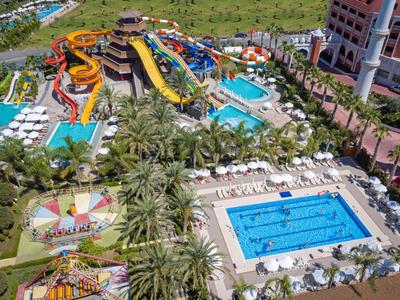 Wasserpark mit Rutschen, Schwimmbecken, Palmen und Liegestühlen in sonniger Umgebung.