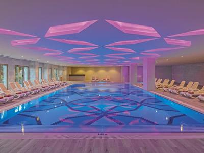 Modernes Hallenbad mit blauer Wasserfläche, vielen Liegestühlen und pink-blau beleuchtetem Deckenmuster.