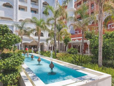 Giardino dell'hotel con una lunga vasca d'acqua rettangolare e fontane decorative circondate da palme.