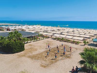 Gruppo che gioca a pallavolo sulla spiaggia con file di ombrelloni e vista sul mare.