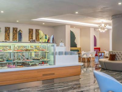 Moderner Cafébereich mit Kuchen-Vitrine, weißen Tischen, Sesseln und heller, eleganter Einrichtung.