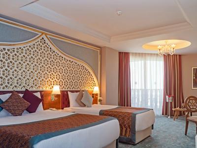 Hotelzimmer mit zwei großen Betten, dekorativem Wandmuster, Lampen und Balkon mit Vorhängen.
