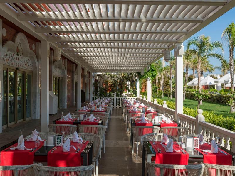 Überdachte Terrasse eines Restaurants mit Tischen und Stühlen, rot dekoriert, neben einem Garten mit Palmen.