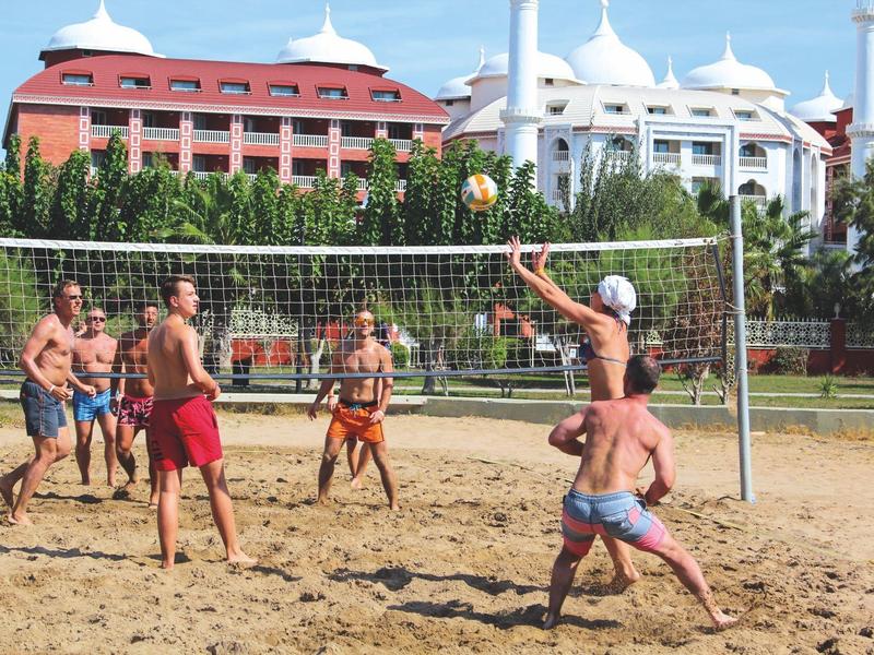 Menschen spielen Volleyball auf einem Sandplatz vor Hotelgebäuden mit weißen und roten Fassaden.