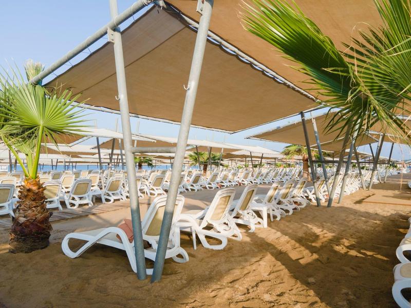 Reihe von weißen Liegestühlen unter einem Sonnenschutz an einem Sandstrand mit Palmen.