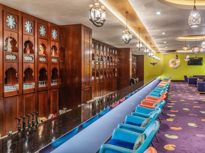 Bunte Bar mit bequemen Stühlen, violettem Teppich und Holzregalen mit Dekorationen.