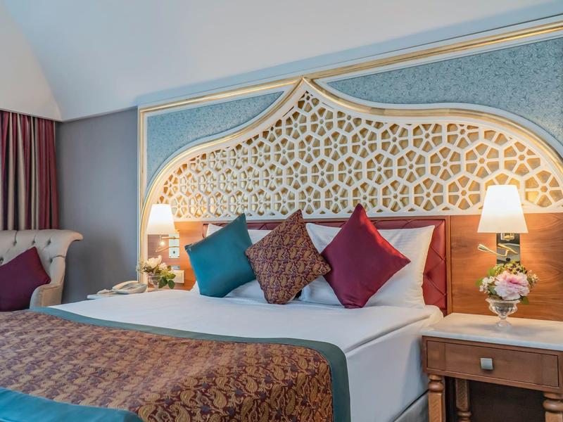 Elegantes Hotelzimmer mit kunstvollem Kopfteil, vier farbigen Kissen und dekorativer Beleuchtung.