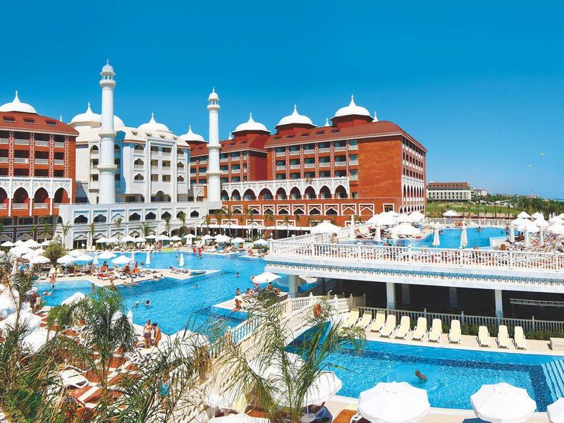 Großes Resort mit roten Dächern, mehreren Pools und blauen Liegestühlen unter blauem Himmel.