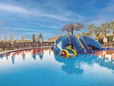 Piscina per bambini con scivolo a forma di pesce colorato e zona relax sotto il cielo azzurro.