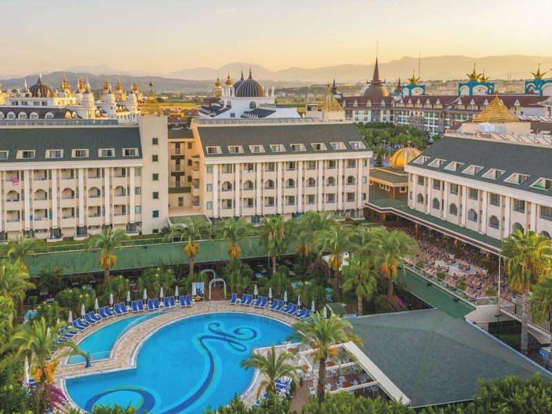 Grande hotel su più piani con piscina curva e palme in un ambiente soleggiato.