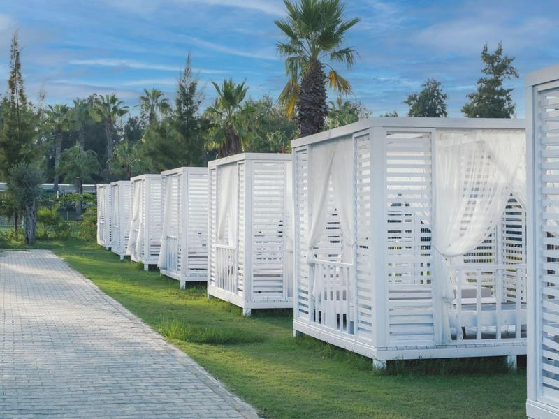 Fila di cabine da spiaggia bianche aperte con tende accanto a un sentiero lastricato e palme.