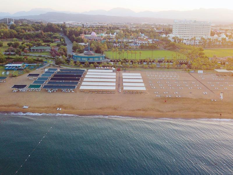 Vista aerea di una spiaggia tranquilla con lettini, edifici e campi verdi sullo sfondo.