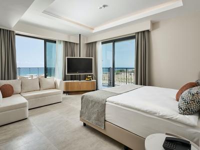 Camera d'hotel luminosa con letto grande, divano, TV e vista mare tramite finestre a tutta altezza.