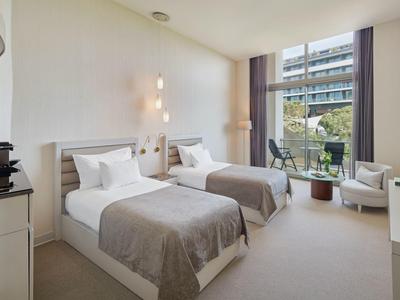 Hotelzimmer mit zwei Einzelbetten, grauen Decken, Stühlen und großer Fensterfront mit Blick nach draußen.