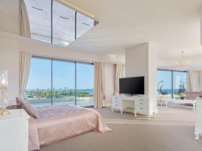 Geräumiges Hotelzimmer mit großem Bett, TV und Panoramafenstern mit Blick auf Wasser.