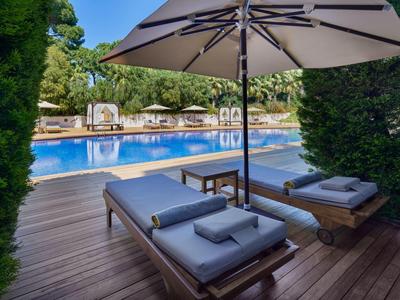 Zwei moderne Liegestühle mit Kissen unter einem großen Sonnenschirm an einem Pool mit Wasser und Bäumen im Hintergrund