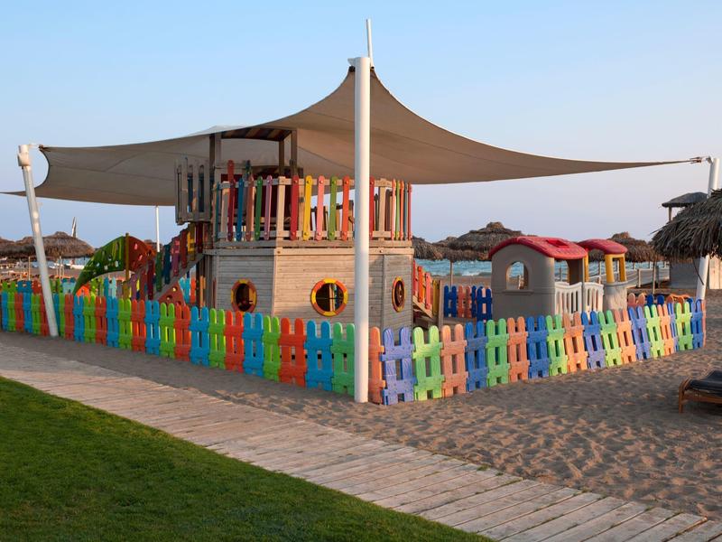 Bunter Kinderspielplatz mit einem überdachten Klettergerüst und Zaun, umgeben von Sand und Rasen.