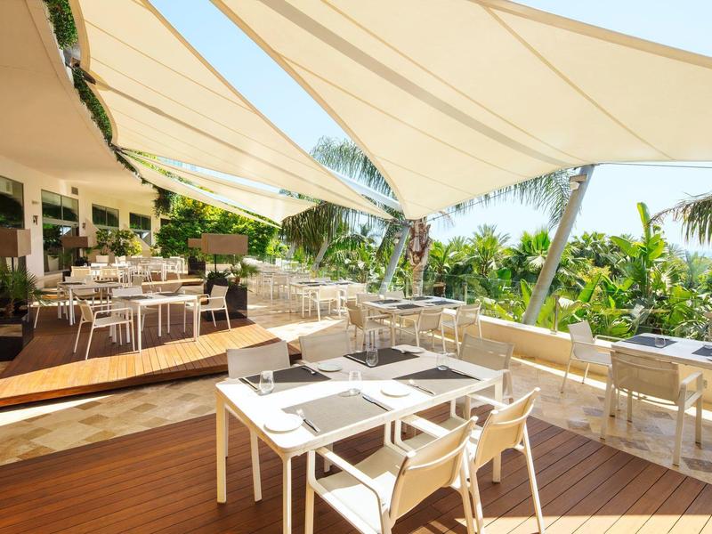 Sonnige Terrasse eines Hotels mit weißen Tischen und Stühlen unter schattenspendenden Segeln