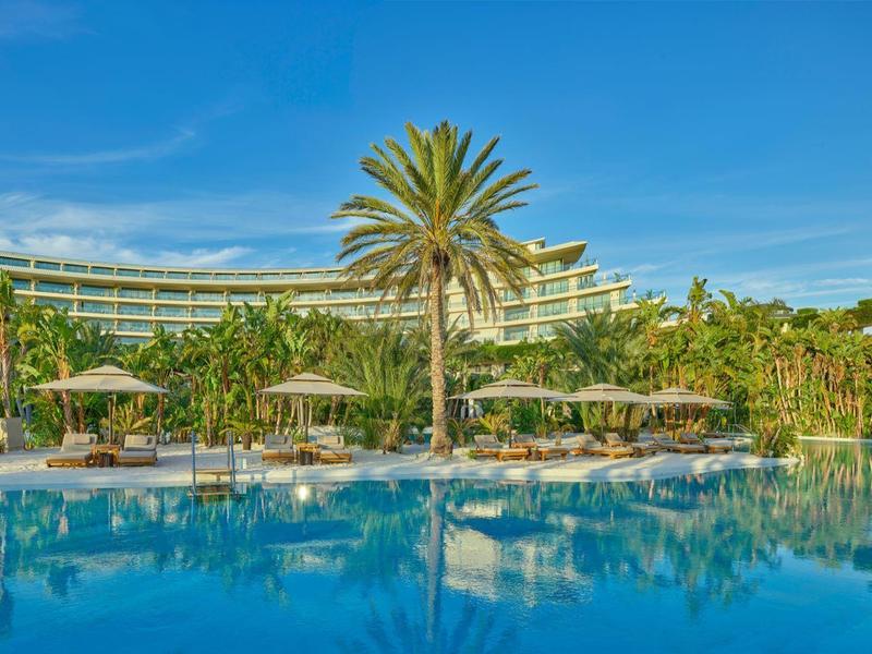 Hotel mit großem Pool, Palmen und Sonnenschirmen unter blauem Himmel