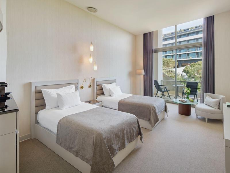 Hotelzimmer mit zwei Einzelbetten, grauen Decken, Stühlen und großer Fensterfront mit Blick nach draußen.