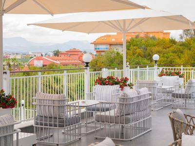 Terrassenbereich mit weißen Tischen und Sitzmöbeln, Sonnenschirmen und Pflanzen mit Ausblick.