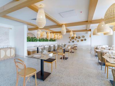 Helles Restaurant mit Holzdeckenbalken, Rattanlampen und Tischen mit weißen Stühlen und Glasvasen.