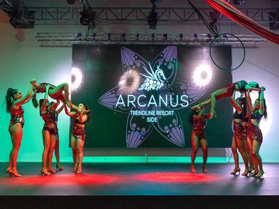 Sechs Tänzerinnen in Bikinis auf Bühne vor LED-Bildschirm mit "Arcanus" Logo und Lichteffekten.