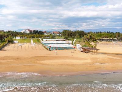 Breiter Sandstrand mit See, Liegestühlen und Palmen vor einem weitläufigen Hotelkomplex.