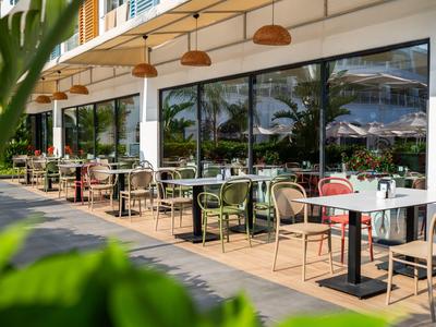 Moderne Außenterrasse eines Hotels mit Tischen, Stühlen und grünen Pflanzen bei Sonnenschein.