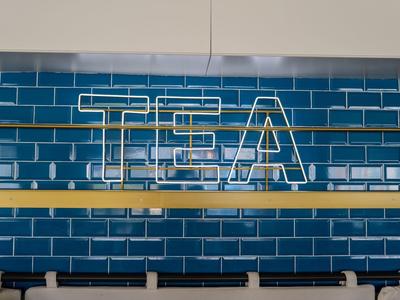 Neonschild mit dem Wort "TEA" vor blau glasierten Kacheln in einer modernen Café-Wand.