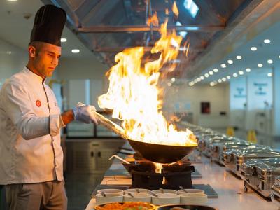 Ein Koch flamt eine Pfanne in einer modernen Hotelküche mit vielen frischen Zutaten auf dem Buffet.