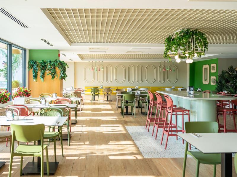Lichte hotel eetzaal met kleurrijke stoelen en grote ramen met planten.