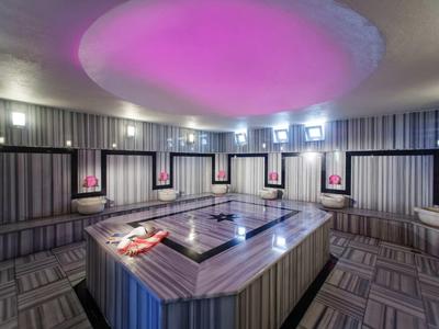 Moderner Wellnessbereich mit beheiztem Whirlpool, Sitzbänken und Decke in hellem Violettlicht.