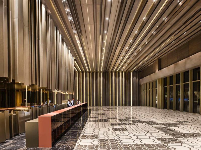 Prostorná hala s mozaikovou podlahou a moderní přepážkou pod osvětleným pruhovaným stropem.