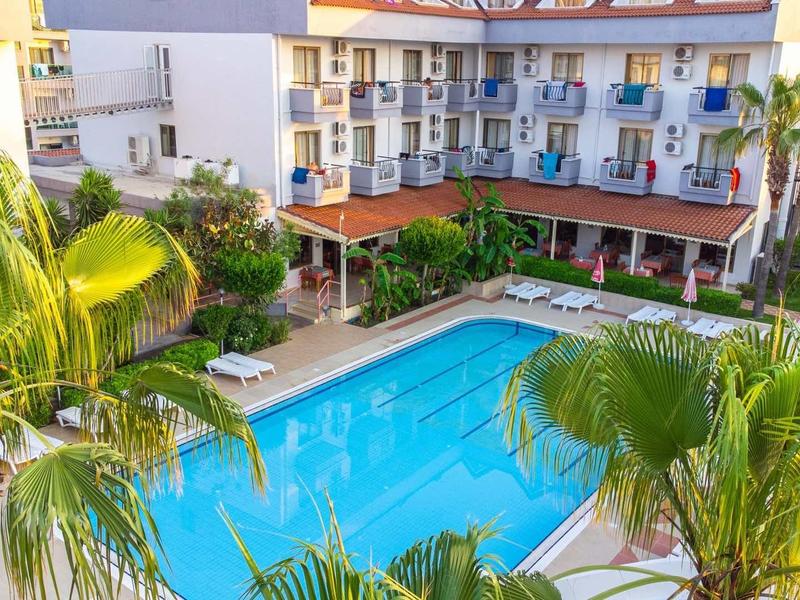 Hotel s velkým obdélníkovým bazénem obklopeným palmami a barevnými balkony.