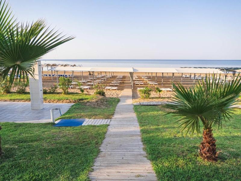 Cestička přes zelenou zahradu s palmami vedoucí k písečné pláži a moři v pozadí.