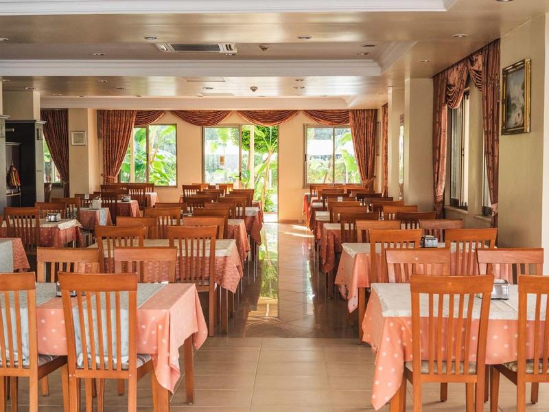 Světlá jídelna s mnoha dřevěnými stoly a židlemi, velká okna vpouštějí denní světlo.