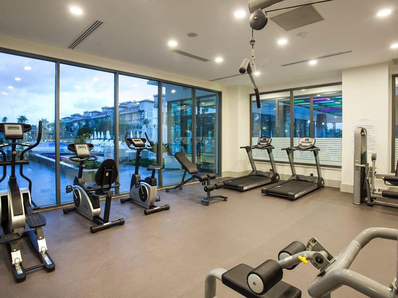 Gimnasio moderno con cintas de correr, bicicletas y grandes ventanas, vista a terraza y paisaje montañoso.