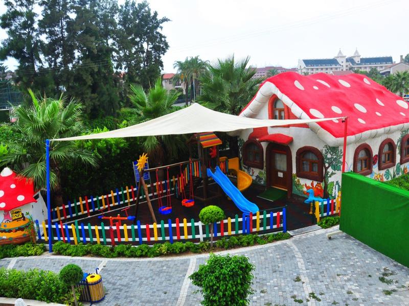 Área de juegos colorida para niños con estructuras de seta y casa, tobogán y cerca, rodeada de plantas.