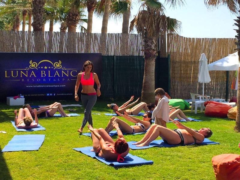 Personas haciendo ejercicio en césped con palmeras y sombrillas al fondo.