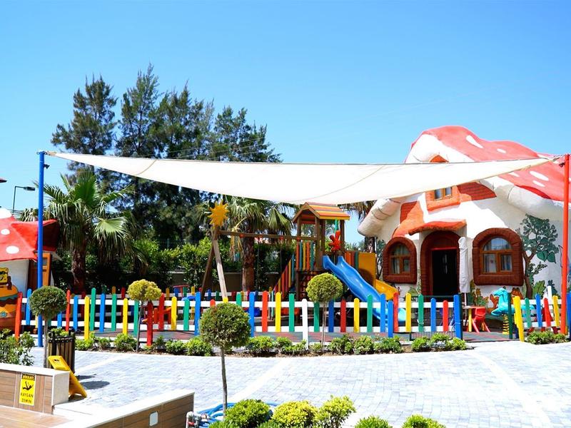 Parque infantil colorido con toldo y cercado multicolor frente a un edificio bajo cielo despejado