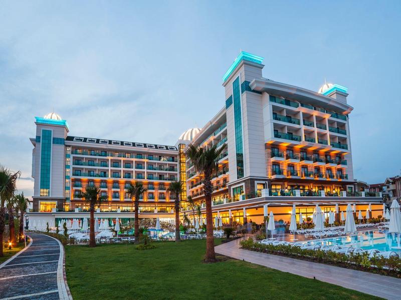 Edificio de hotel moderno con balcones iluminados y piscina en un entorno tropical al atardecer.