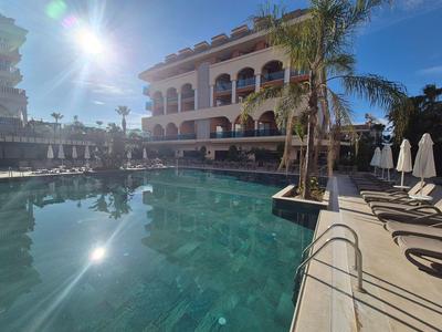 Bâtiment d'hôtel avec piscine, palmiers et chaises longues par temps ensoleillé.