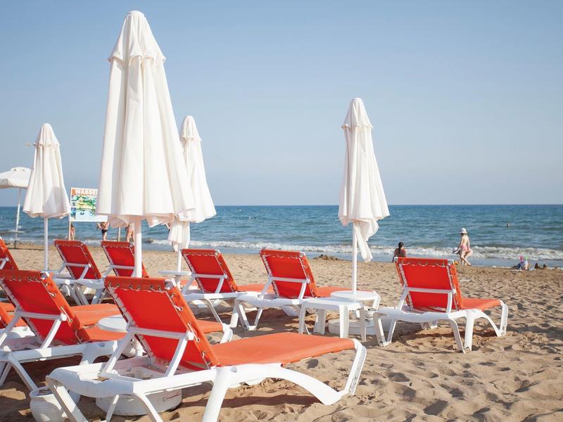 Red sun loungers and closed white umbrellas on a sandy beach with the sea in the background.