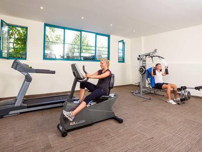 Ein heller Fitnessraum mit zwei Personen auf Trainingsgeräten vor großen Fenstern und heller Wand.