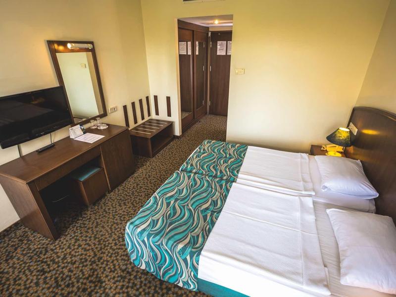 Pokój hotelowy z łóżkiem podwójnym, telewizorem, biurkiem i lustrem w ciepłych kolorach.