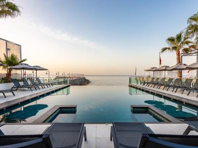 Infinity-Pool mit Liegestühlen und Palmen, ruhiges Meer im Hintergrund bei klarem Himmel.